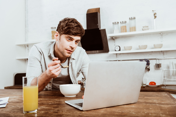 male freelancer eating corn flakes and working on laptop at table in kitchen - Фото, зображення