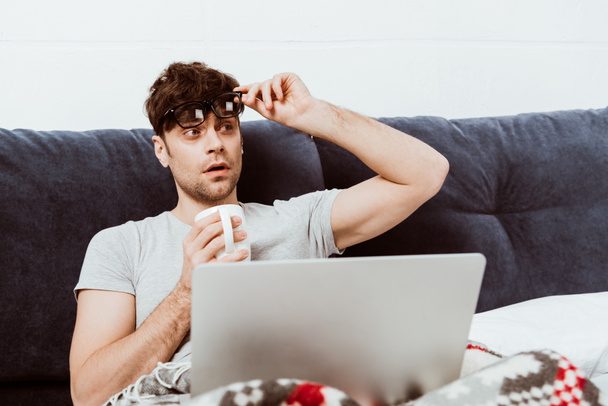 young male freelancer taking off eyeglasses and holding coffee cup on bed with laptop at home - 写真・画像