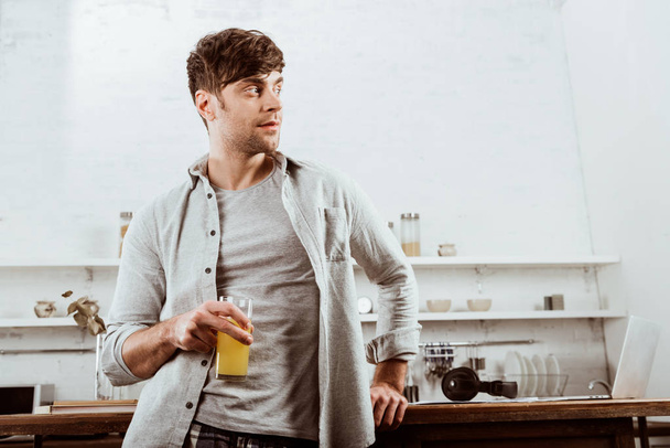 ラップトップでのキッチン テーブルの近くのオレンジ ジュースの立っている若い男性フリーランサー  - 写真・画像