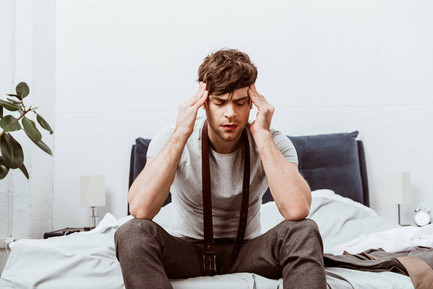 joven hombre de negocios con cinturón sobre el cuello sentado en la cama y con dolor de cabeza en casa
 - Foto, imagen