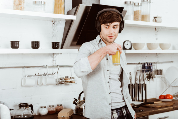jongeman hoofdtelefoon drinken sinaasappelsap op ontbijt in keuken thuis - Foto, afbeelding