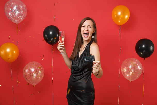 Szczęśliwa młoda kobieta w małej czarnej, krzycząc docenianie, posiadania karty kredytowej kieliszek szampana na jasnym tle czerwonych balonów. Nowy rok, urodziny makieta holiday party koncepcja - Zdjęcie, obraz
