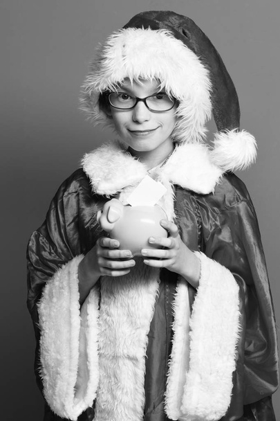 young cute santa claus boy with glasses in xmas sweater and new year christmas hat holding pink piggy pig bank on red studio backgroun - Φωτογραφία, εικόνα