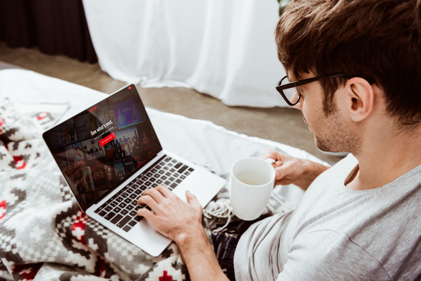 vista lateral del hombre sosteniendo la taza de café y utilizando el ordenador portátil con netflix en la pantalla en la cama en casa
 - Foto, Imagen