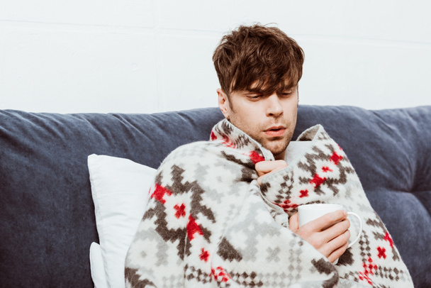 junger kranker Mann in Decke gehüllt trinkt zu Hause Tee im Bett  - Foto, Bild