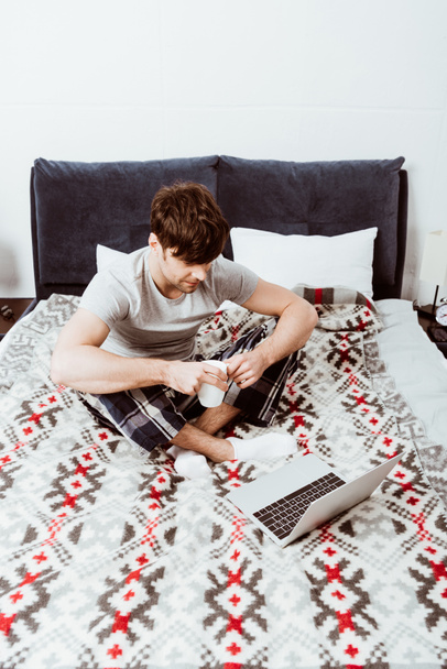 young male freelancer with coffee cup sitting on bed with laptop at home - Foto, Imagen