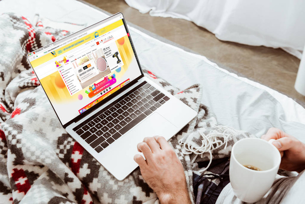 cropped image of man holding coffee cup and using laptop with aliexpress on screen in bed at home - Zdjęcie, obraz