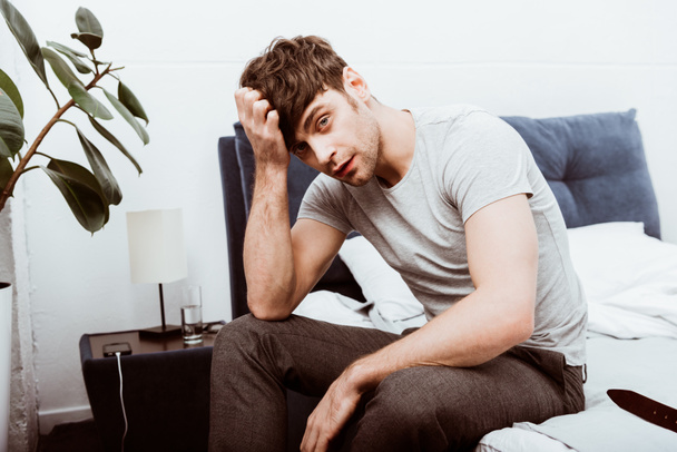 unavený mladý podnikatel při pohledu na fotoaparát a sedí na posteli doma - Fotografie, Obrázek