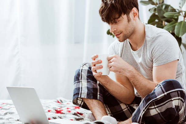 bell'uomo freelance con tazza di caffè seduto sul letto con computer portatile a casa
 - Foto, immagini