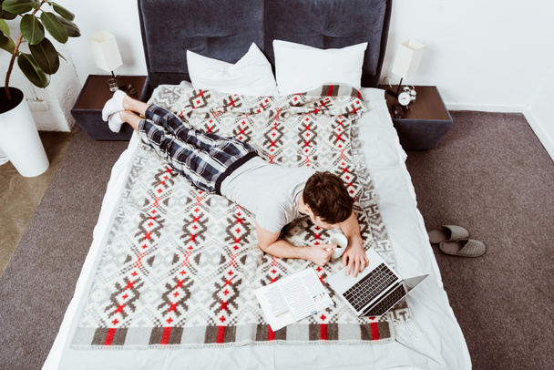 hoge hoekmening van mannelijke freelancer met koffiekopje tot op bed met laptop thuis - Foto, afbeelding