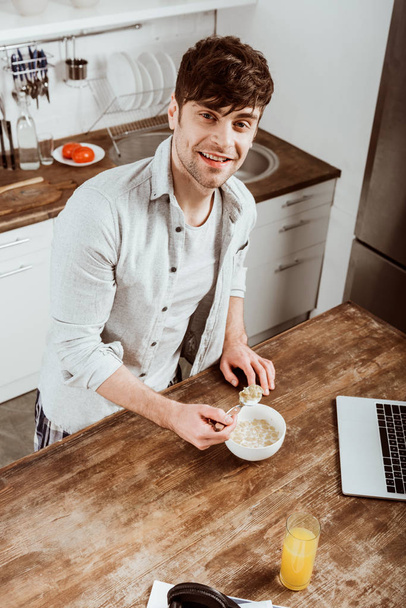 vista de ángulo alto del freelancer masculino comiendo hojuelas de maíz en el desayuno en la mesa con computadora portátil en la cocina en casa
 - Foto, Imagen