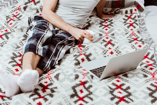 частковий вид чоловіка фрілансера з чашкою кави, що сидить на ліжку з ноутбуком вдома
 - Фото, зображення