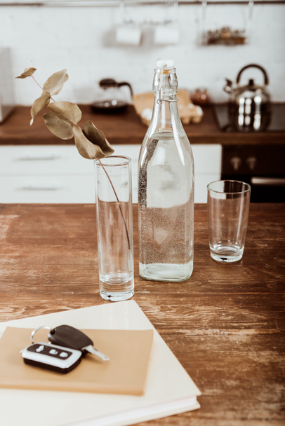 крупним планом вид на віддалений ключ автомобіля, пляшка води і гілка у вазі за дерев'яним столом на кухні
  - Фото, зображення