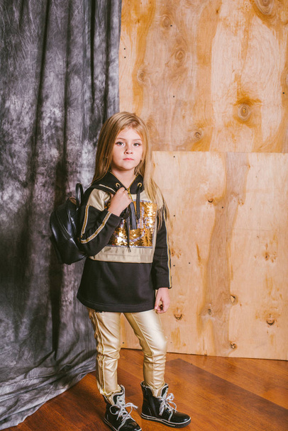 Little cute girl with blond hair in fashionable leggings, with gold and sequins, with a backpack - Foto, imagen