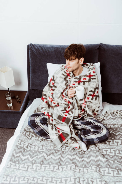 hoge hoekmening van zieke man gewikkeld in een deken zitten met een kopje thee op bed thuis  - Foto, afbeelding