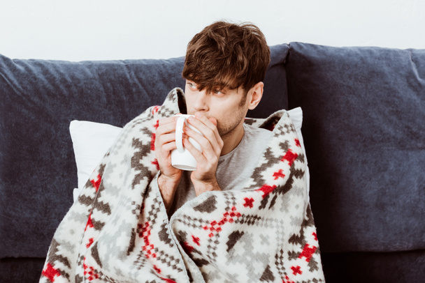 selective focus of sick man wrapped in blanket drinking tea in bed at home  - Foto, Bild