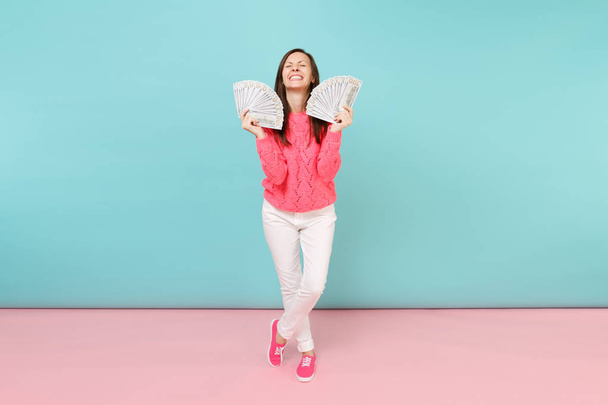 Volledige lengte portret opgewonden vrouw in gebreide roze trui, witte broek houden veel dollars bankbiljetten geïsoleerd op helder roze pastel blauwe muur achtergrond in de studio. Geld concept. Bespotten kopie databaseruimte - Foto, afbeelding