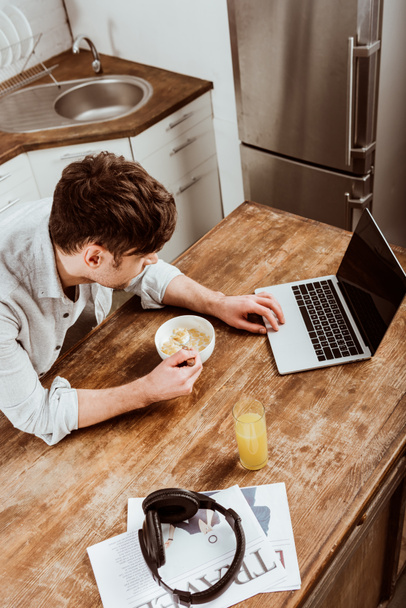 自宅の台所でノート パソコンとテーブルで朝食にコーンフレークを食べる男性のフリーランサーのハイアングル - 写真・画像