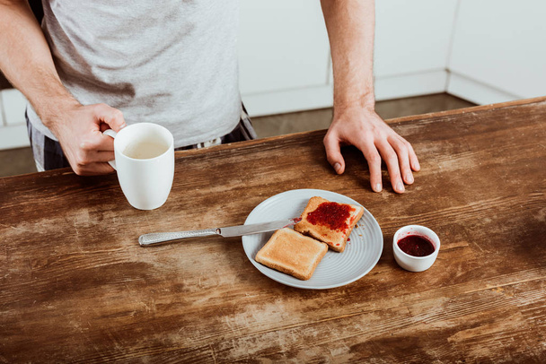 cropped image of man with coffee cup standing at table with toasts and jam in kitchen - Φωτογραφία, εικόνα