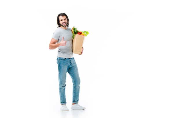 Handsome man keeping grocery bag,  showing thumb up and smiling at camera isolated on white - Photo, Image
