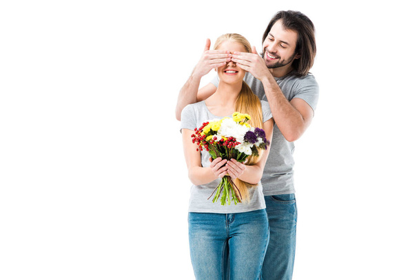 ehrfürchtige Frau mit Blumen, während Mann ihre Augen schließt, um eine Überraschung zu machen, isoliert auf weiß - Foto, Bild