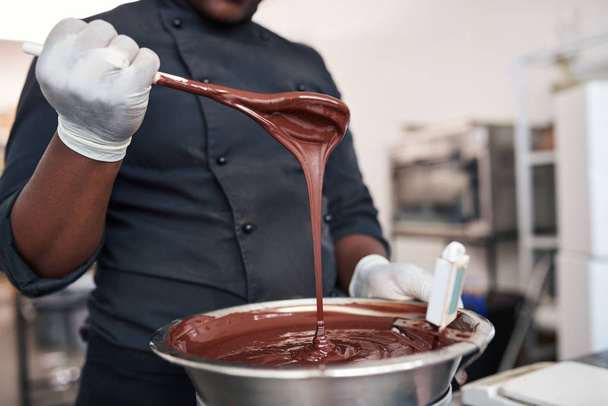 Closeup of a worker holding a spoon full of melted chocolate while preparing ingredients in an artisanal chocolate making factory - Foto, immagini
