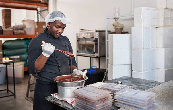 Worker standing at a table in an artisanal chocolate making factory mixing melted chocolate in bain marie with a spoon - Foto, afbeelding