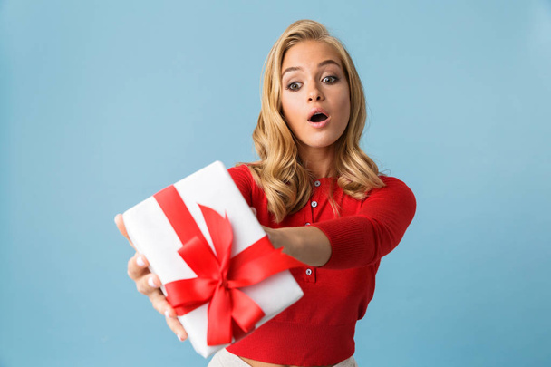 Portrait of joyous blond woman 20s wearing red shirt holding gift box isolated over blue background in studio - Photo, Image