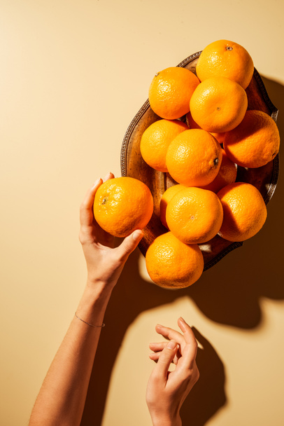 Cropped shot of woman holding tangerine on beige background with metal bowl - Foto, Bild