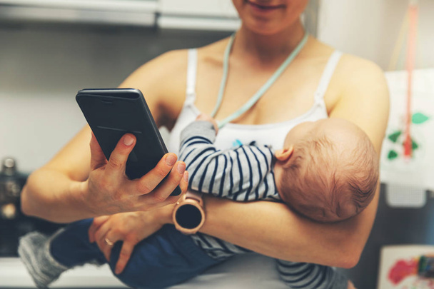 äiti käyttää älypuhelinta pitäen vauvaa
 - Valokuva, kuva