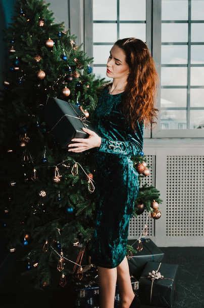 Pretty girl holding a gift box near Christmas tree, windows in the background. New Year and Christmas concept. Home and family warmth - 写真・画像