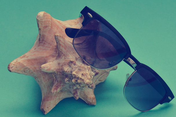 Decorative seashell and sunglasses on a blue paper background. The concept of a resort holiday. - Foto, imagen