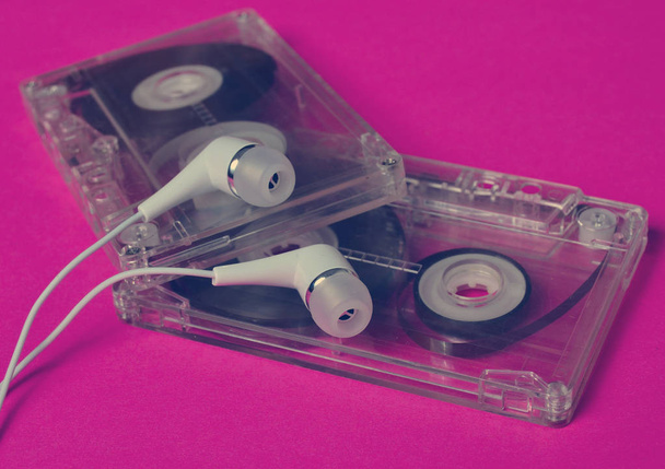 Ретро-технологія. Пластикова прозора аудіокасета та білі вакуумні навушники на яскраво-рожевому фоні. 80-ті роки
. - Фото, зображення