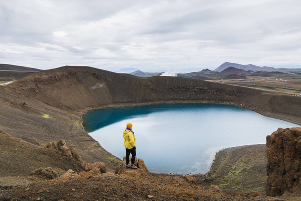 Frau in gelbem Regenmantel steht im wassergefüllten Krater des Vulkans Krafla in Island. Panoramaaufnahme, bewölkter Tag. - Foto, Bild