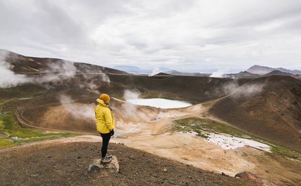Nő a sárga esőkabátot állt a víz tele kráter a Krafla vulkán, Izland. Panorámás lövés, felhős nap. - Fotó, kép