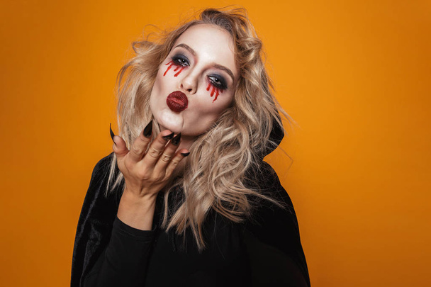 Frightening woman wearing black costume and halloween makeup looking at the camera isolated over yellow background - Foto, immagini