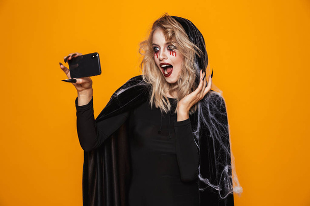 Beautiful woman wearing black costume and halloween makeup taking selfie photo on mobile phone isolated over yellow background - Foto, immagini