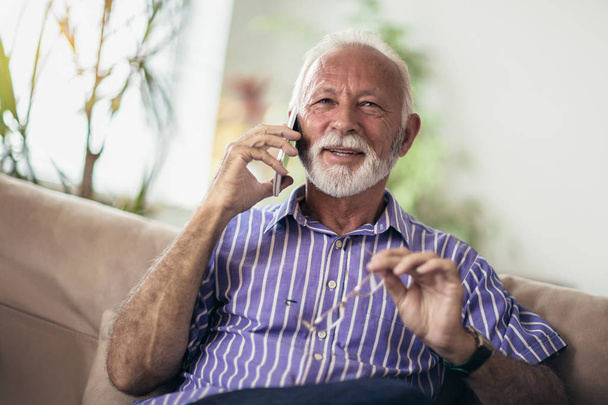 Старший мужчина болтает по мобильному телефону, расслабляясь на диване в своей гостиной
 - Фото, изображение