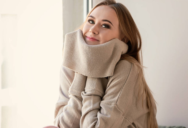Beautiful young woman in warm sweater sitting near window at home - Foto, afbeelding