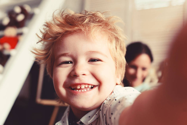 Boy spends fun time in playroom. Child with cheerful face and blond hair, close up. Joy and games concept. Kid with his mother on light room background, defocused - Photo, image