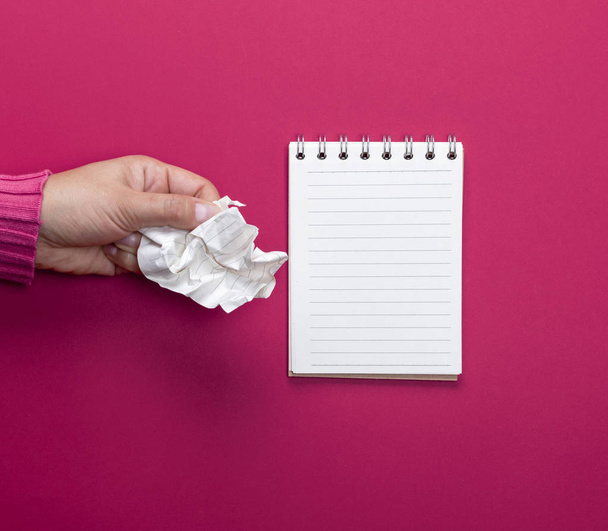 mano femenina sosteniendo una hoja de papel blanco arrugado y un cuaderno abierto sobre un fondo rosa
 - Foto, Imagen
