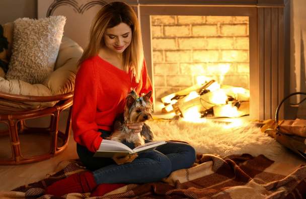 Woman with cute dog reading book near fireplace - Фото, изображение