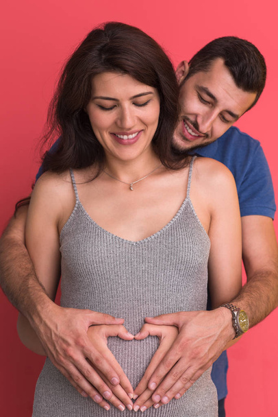 Happy young couple showing heart with their hands on belly of pregnant wife isolated on red background - Foto, Bild