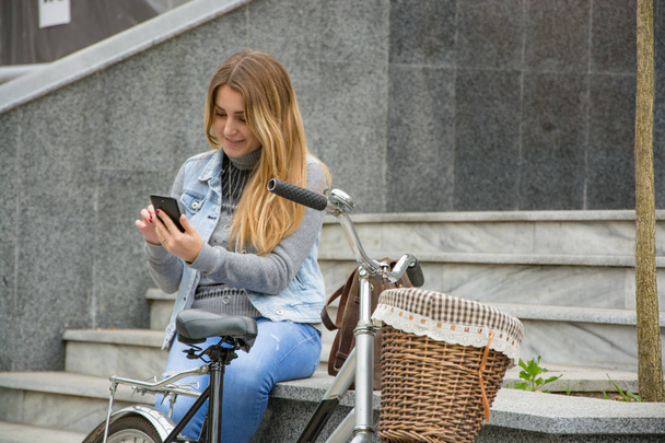 νεαρή γυναίκα αναβάτη ποδήλατο χρήση cellphone, αστικό φόντο - Φωτογραφία, εικόνα