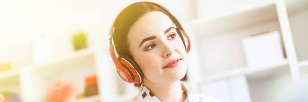 Beautiful young girl sits in headphones at desk in office, holds a notebook in her hand and prints on the keyboard. Photo with depth of field, focus on girl. - 写真・画像