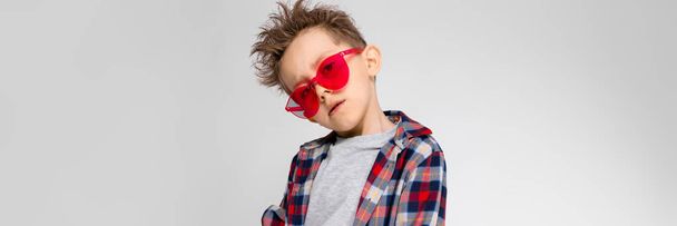 Ένα όμορφο αγόρι σε ένα καρό πουκάμισο, γκρι πουκάμισο και τζιν στέκεται σε γκρι φόντο. Ένα αγόρι σε κόκκινα γυαλιά ηλίου. Το αγόρι που το χέρι στο μπράτσο του. - Φωτογραφία, εικόνα