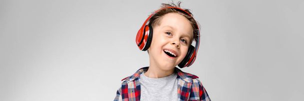 A handsome boy in a plaid shirt, gray shirt and jeans stands on a gray background. A boy in red headphones. The boy holds his hands on his stomach. The boy laughs. - Photo, image