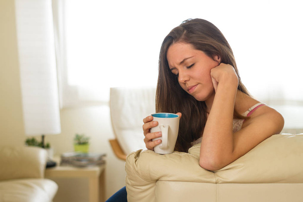 Miserable sad young brunette girl who is feeling bad while taking a cup of hot tea or coffee in the living room of her brightly lit house - Foto, Imagen