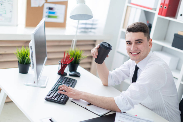 Un joven sentado en una mesa en la oficina y sosteniendo un vaso de café y escribiendo en el teclado
. - Foto, Imagen