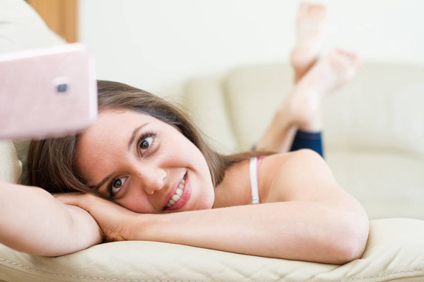Pretty young brunette caucasian woman resting on the couch while making a selfie with surprised surprise face and face of surprise and uncertainty, with legs crossed - Valokuva, kuva